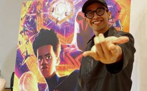 A person wearing a dark shirt, glasses, and a cap smiles while making a distinct hand gesture towards the camera. Behind them is a large, colorful poster of the animated movie "Spider-Man: Into the Spider-Verse," featuring various Spider-Man characters and Netflix branding. CodersLink 2024.