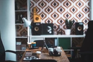 A cozy, cluttered office space with a wooden desk featuring a coffee cup, gadgets, a laptop, and headphones. Behind the desk is a shelf with speakers, a plant, and other decorative items. The patterned wallpaper adds a retro touch to the room—perfect for nearsourcing recruitment projects in Latin America. CodersLink 2024.