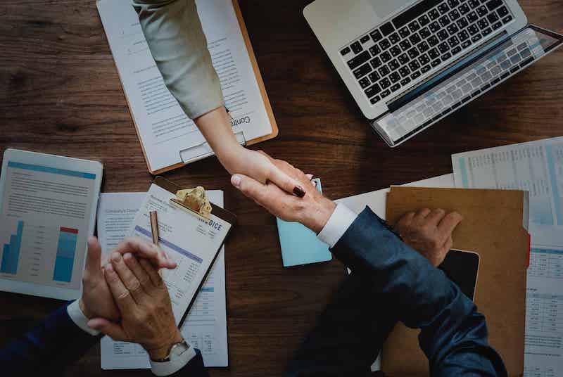 Overhead view of a business meeting with people shaking hands. The table is cluttered with documents, a clipboard, a laptop, and pens. One person reaches out to shake hands with another, indicating agreement or conclusion of a discussion on remote work policies. CodersLink 2024.