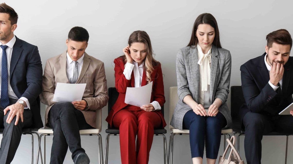 Five individuals sit in a row of chairs, seemingly in a waiting area. They are dressed in business attire and appear focused, reading documents or looking thoughtful. The plain, light-colored wall serves as the backdrop for these job seekers eager to conseguir empleo en TI through CodersLink. CodersLink 2024.